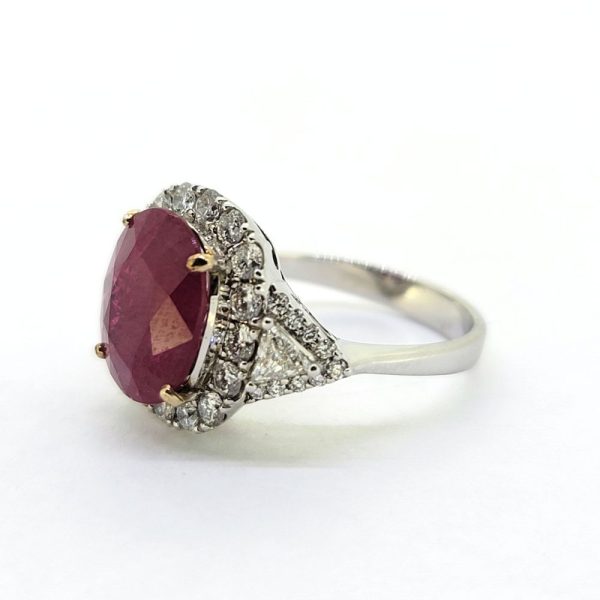 Contemporary 4.79ct Ruby and Diamond Cluster Engagement Ring, 4.79ct oval red ruby flanked by trillion diamonds all within a brilliant-cut diamond halo border in 18ct white gold