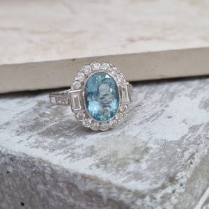 A vintage inspired ring with a 2.10 carat oval aquamarine in the centre surrounded by 0.55 carats of diamonds, rubover set in 18ct white gold.