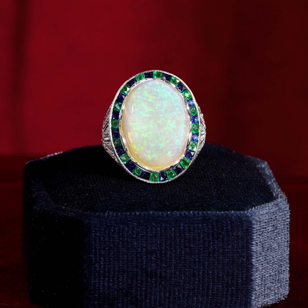 8.73ct Opal Emerald Sapphire Cluster Cocktail Ring, central luminous opal encircled by vivid French-cut emeralds alternated with blue sapphires in 18ct white gold with diamond shoulders
