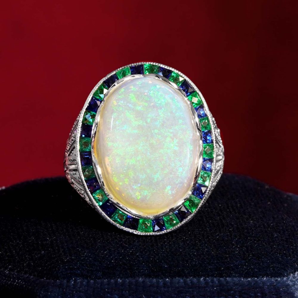 8.73ct Opal Emerald Sapphire Cluster Cocktail Ring, central luminous opal encircled by vivid French-cut emeralds alternated with blue sapphires in 18ct white gold with diamond shoulders